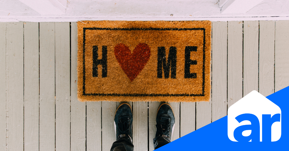 doormat on a wooden deck with the word home on in with a heart in the o and a person's feet in front of the mat and an ar symbol on the right