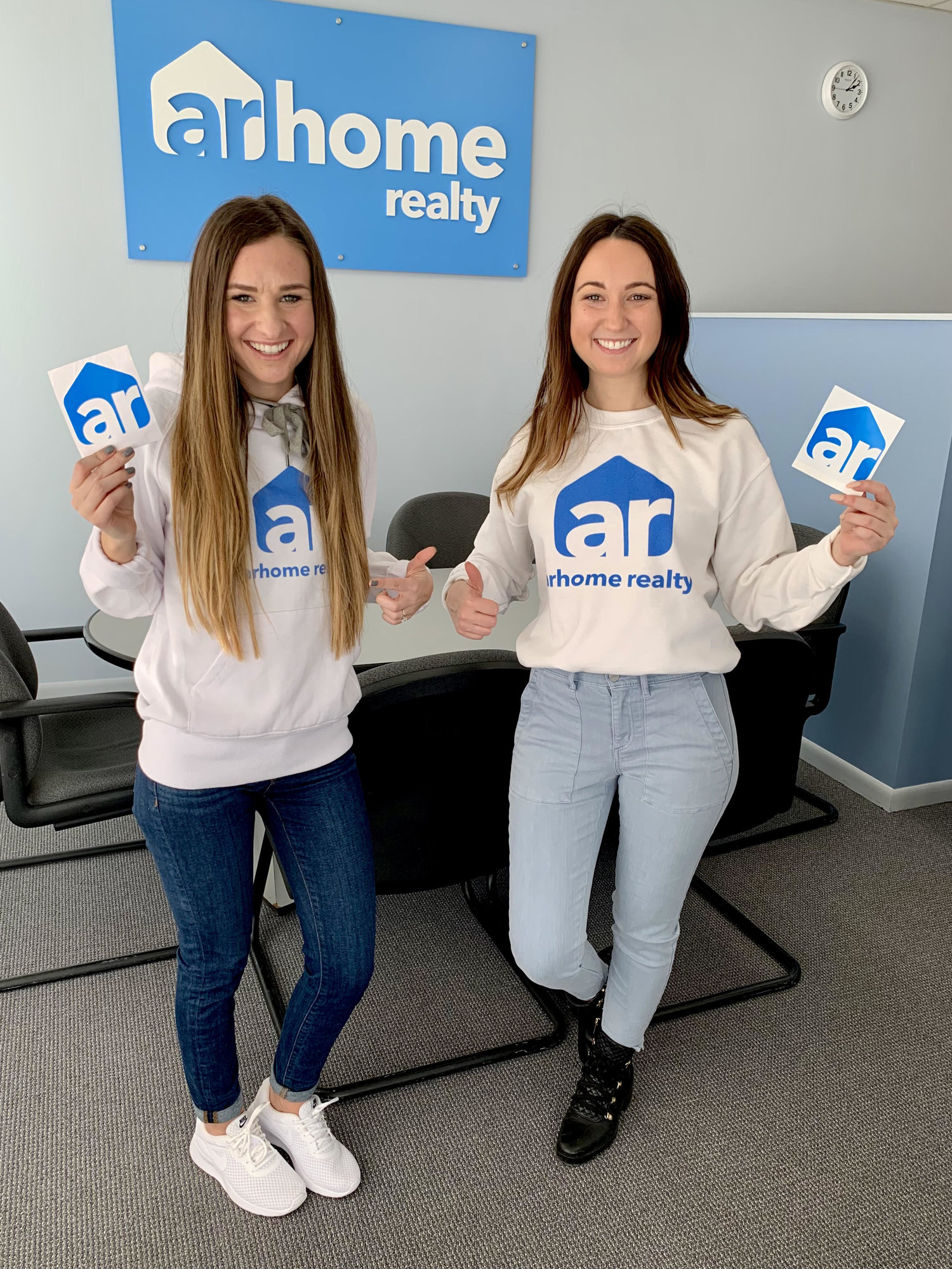 two women standing in ar logo sweaters with ar logo stickers in front of the arhome realty sign